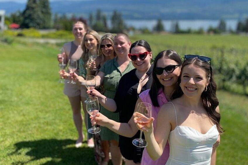 A group of people holding wine glasses in a grassy field with a body of water and trees in the background. 4