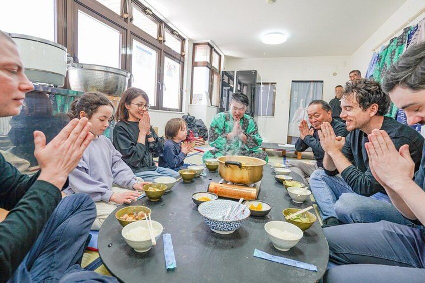Een groep mensen zit rond een tafel met schalen eten, sommigen gebruiken stokjes, in een ruimte met grote ramen. 2