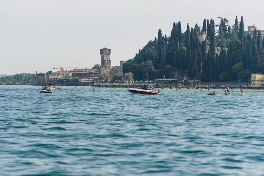 A calm body of water with boats, a tower, and a dense tree line in the background. 1