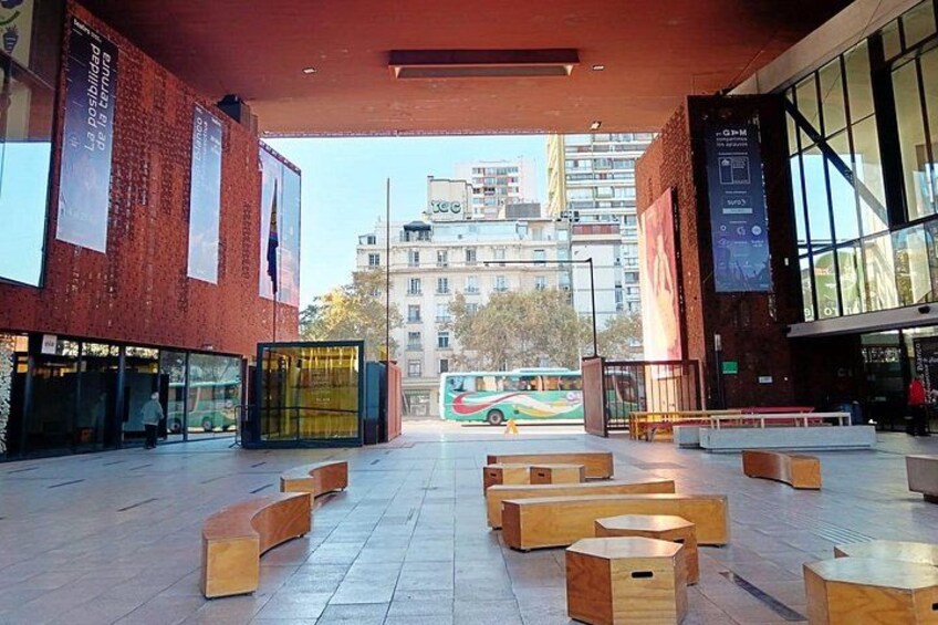Un edificio moderno con amplias ventanas y fachada de ladrillo. Se observan bancos de madera y un autobús al fondo. 2