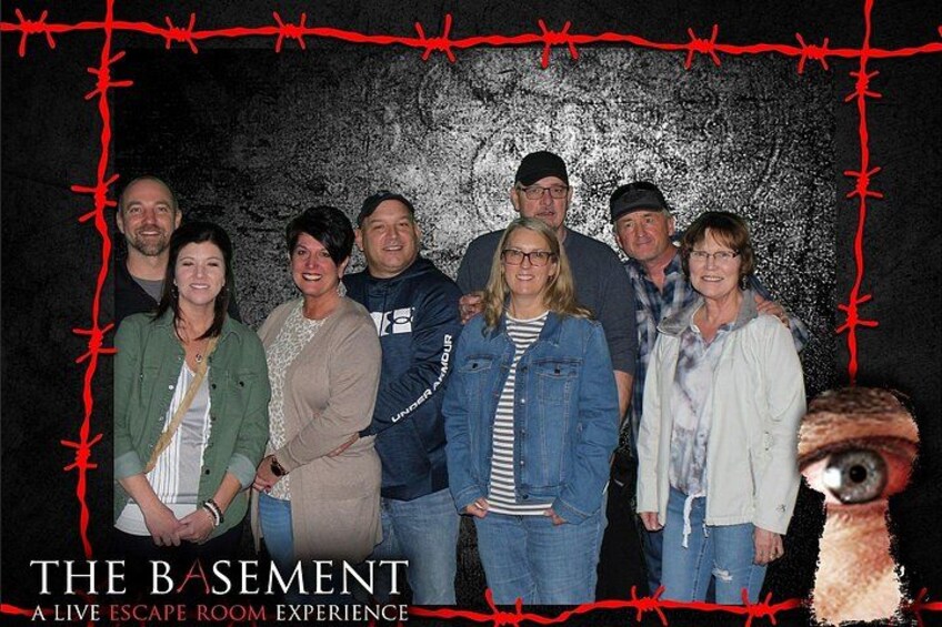 A group of people in front of a wall with a barbed wire design, titled 'THE BASEMENT A LIVE ESCAPE ROOM EXPERIENCE'. 3