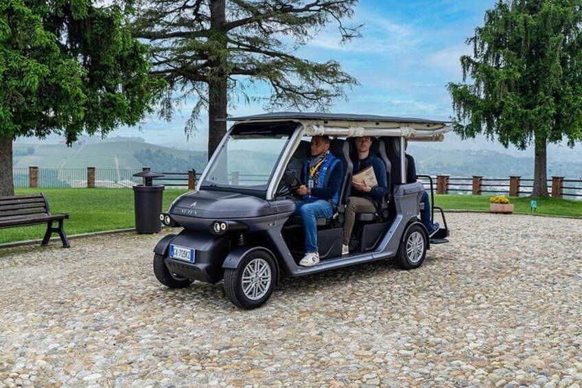 A golf cart with two passengers on a gravel path with trees and a bench. 4