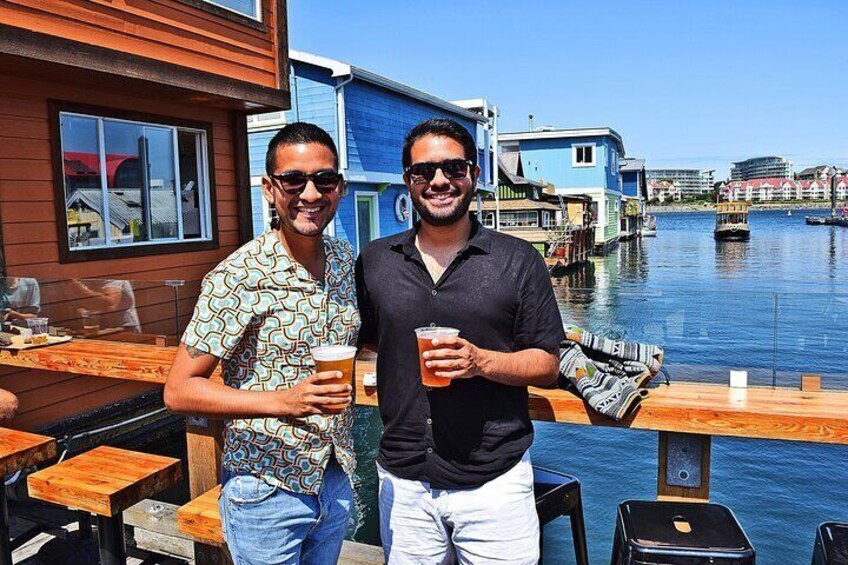 Twee personen staan bij een waterfront bar met houten tafels en stoelen, terwijl ze iets drinken. 1