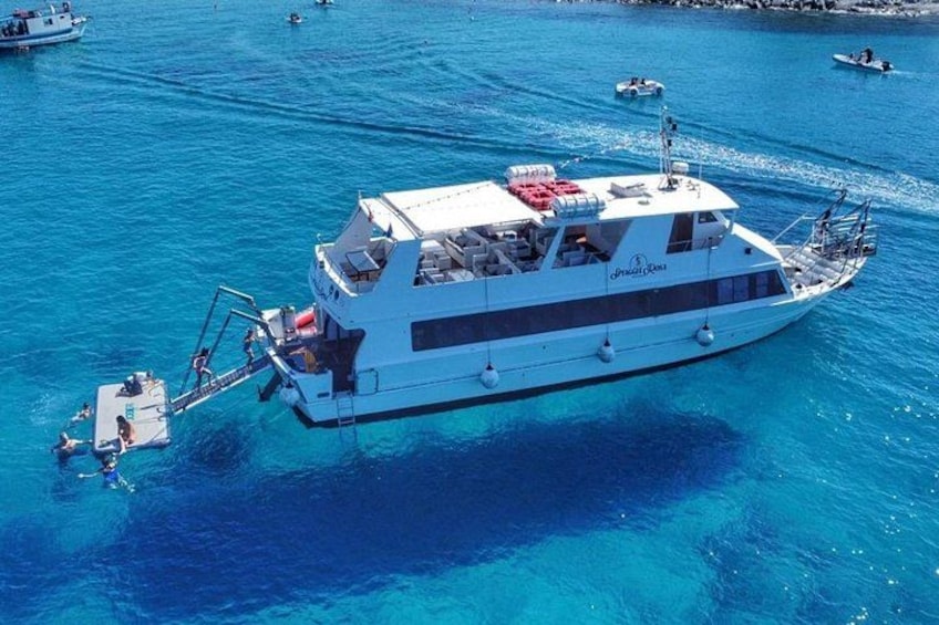 A large white boat with a red deck and a logo on the side, floating on clear blue water with other smaller boats in the background. 5