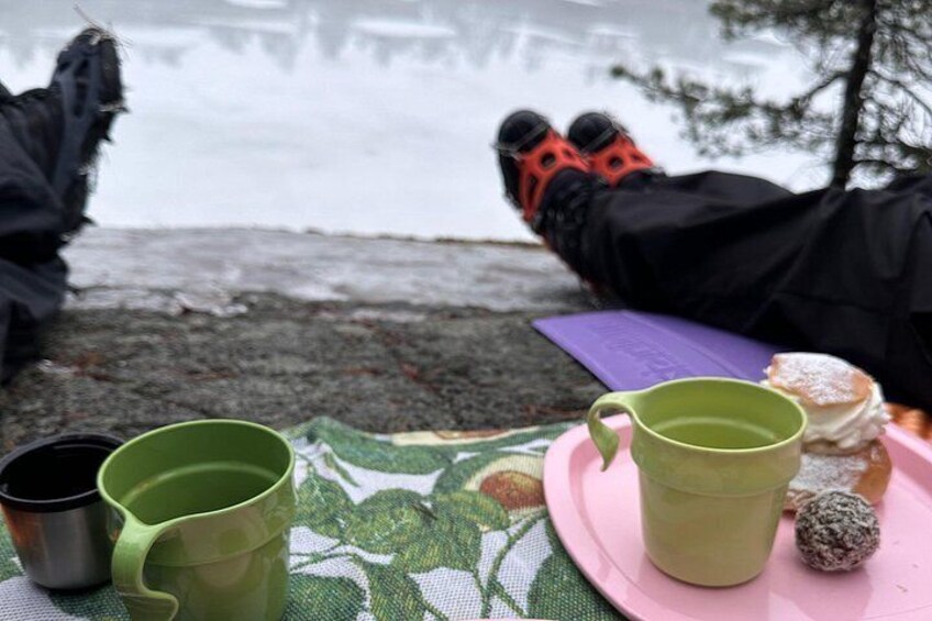 A person lying on a blanket by a lake, with a thermos, mug, and donut on a plate. 3