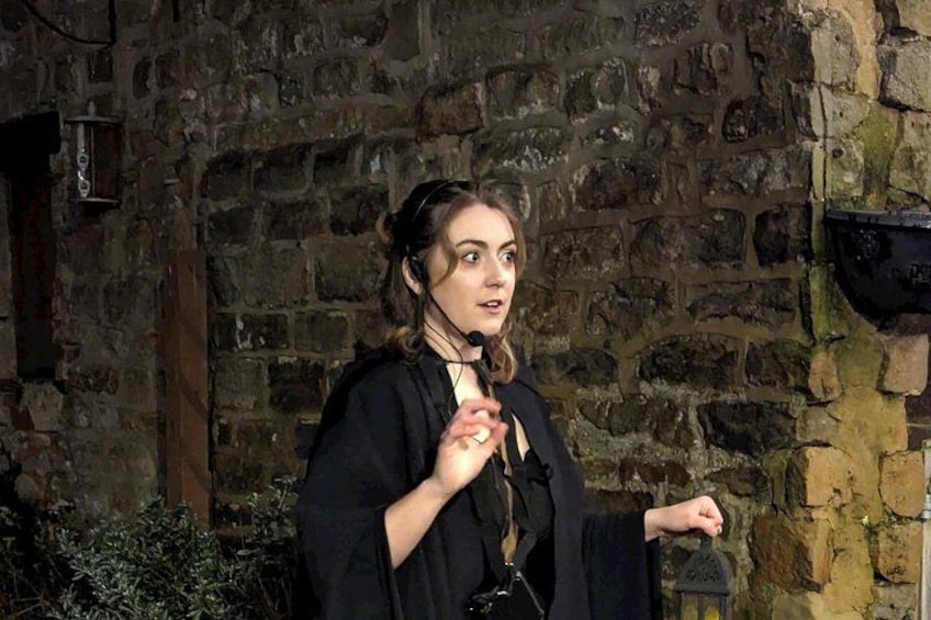 A person in black attire standing in front of a stone wall with a lantern. 2