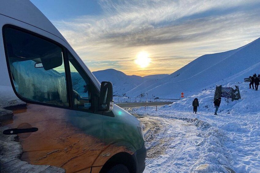 A snowy landscape with a vehicle parked on a road, mountains in the background, and a sunset. 2