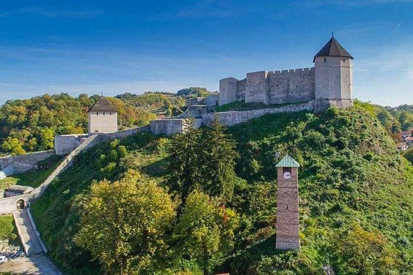 A medieval castle on a hill with a watchtower, surrounded by greenery and a clear sky. 3