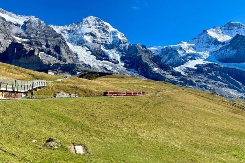 A red train on a track, a building, and snow-capped mountains. 1
