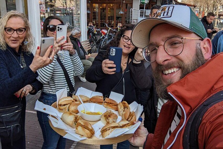 People holding food items, likely sandwiches, outdoors with a building in the background. 1