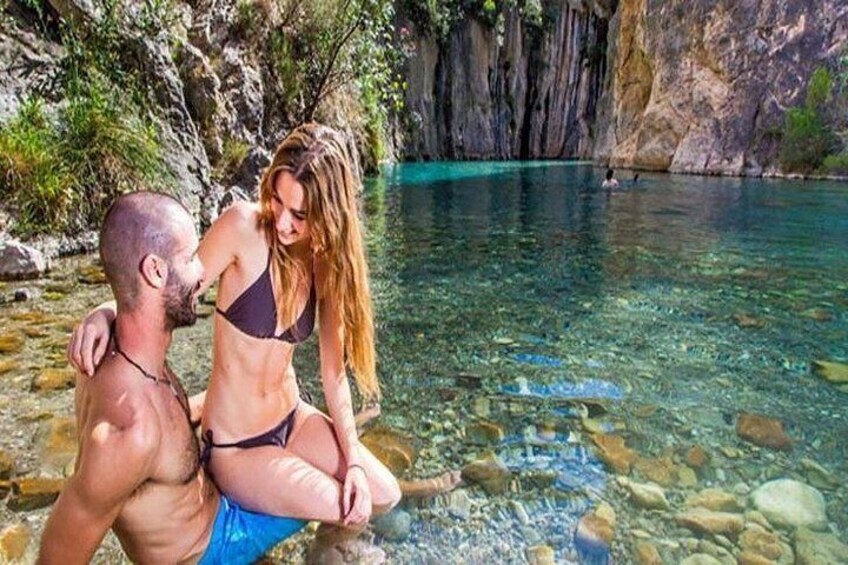 A couple sitting by a clear, rocky pool surrounded by cliffs. 2