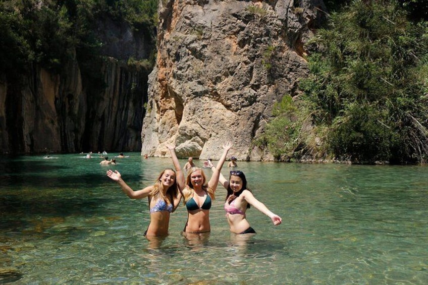 Three people in bikinis standing in clear water with a rocky cliff and greenery in the background. 2