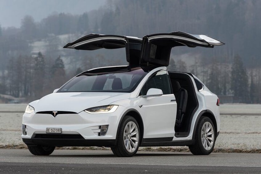 A white Tesla with its trunk open, parked on a road with a snowy landscape in the background. 2