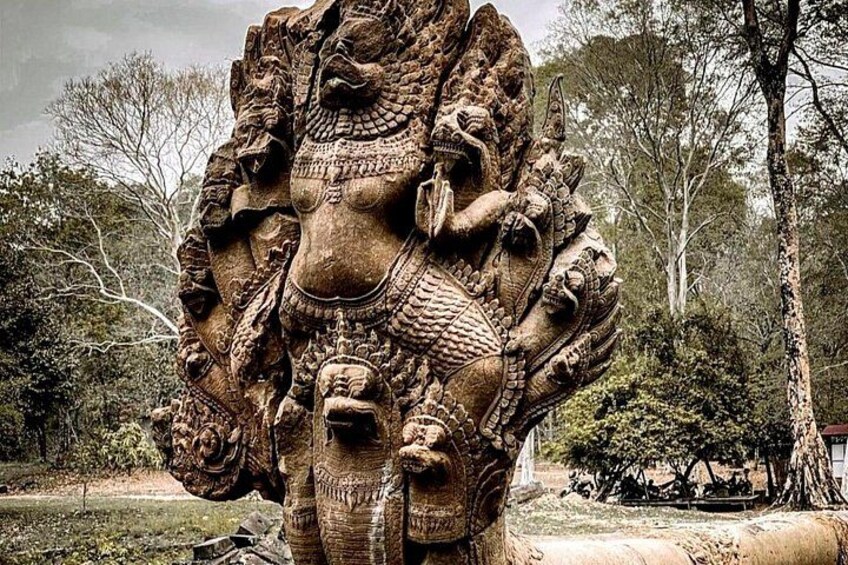 Une grande sculpture en pierre, dotée de multiples visages et de motifs complexes, se dressant sur un fond de feuillage et d’un ciel nuageux. 2