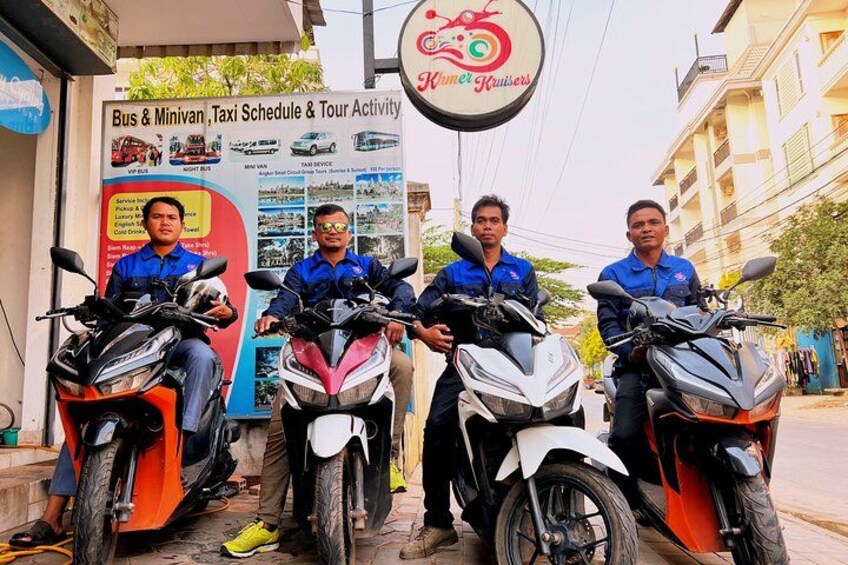 Trois hommes sur des scooters devant un panneau publicitaire annonçant des services d’autobus et de navettes. 1