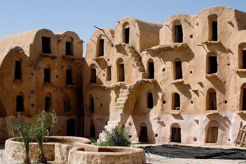 A traditional mud-brick building with multiple arched windows and doorways, surrounded by palm trees and a dry, barren landscape. 2