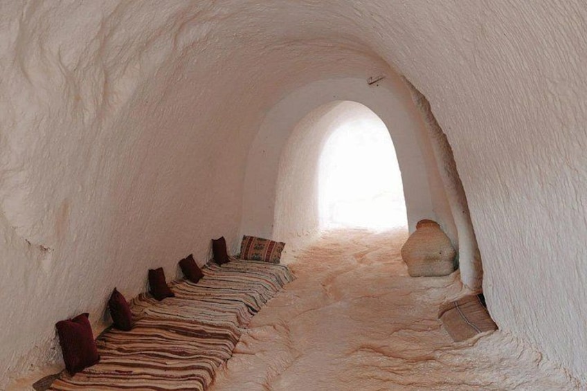 An underground room with a sloped ceiling, a stone bench, and a patterned rug. 4