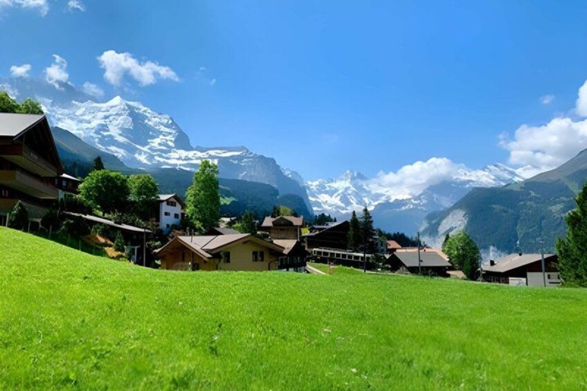 A green hillside with buildings, a clear blue sky, and snow-capped mountains in the background. 3