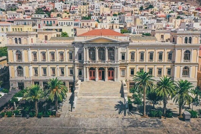 A large classical building with a grand staircase leading to a red door, surrounded by palm trees and a densely packed urban area. 1