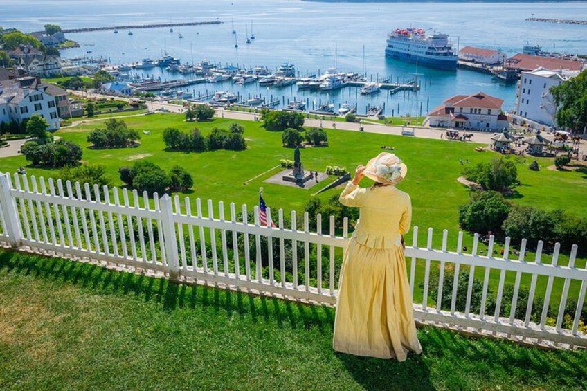 A woman in a yellow dress standing by a white picket fence overlooking a marina with boats and a cruise ship. 5