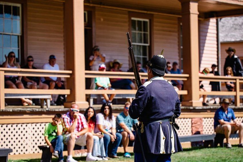 A historical reenactment scene with a uniformed individual in the foreground, spectators seated on bleachers in the background. 2