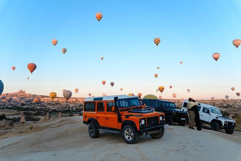 Paisaje desértico con una caravana de vehículos y globos aerostáticos al fondo. 3