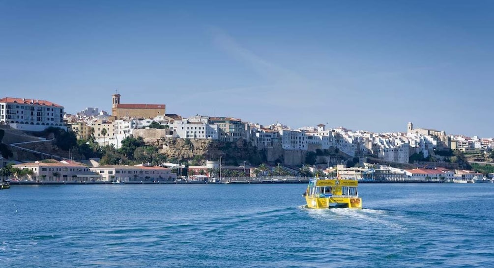 A coastal cityscape with a yellow boat on the water, buildings on a hillside, and a clear sky. 1