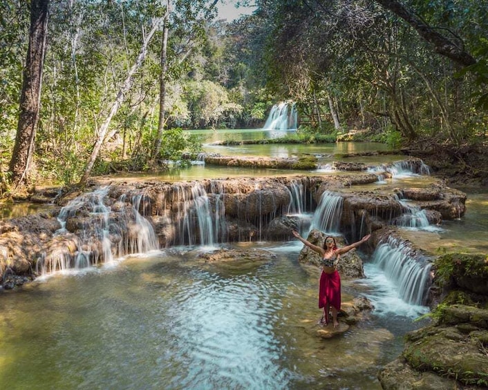 A waterfall, a person standing on rocks, and a forested area. 1