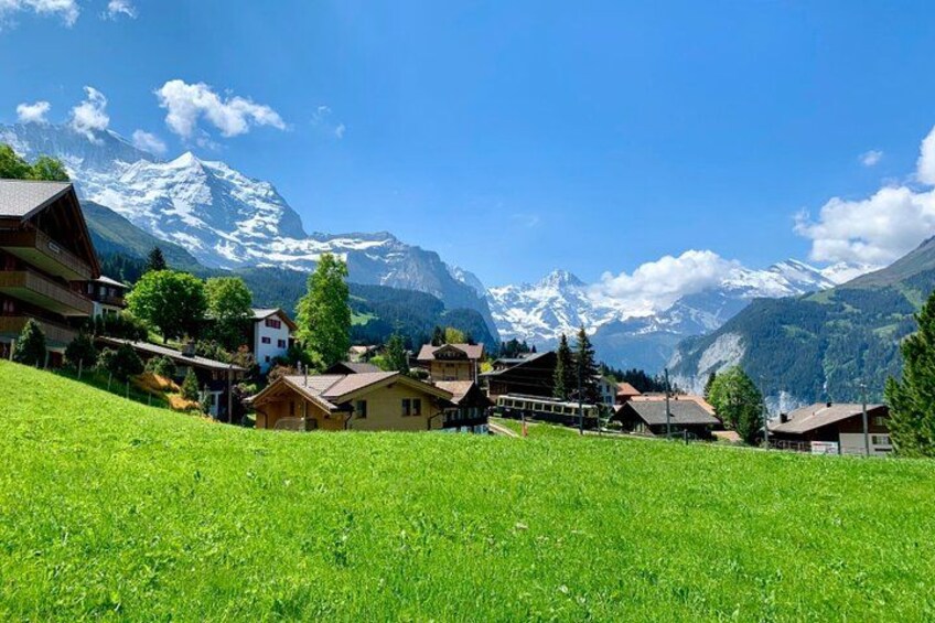 A village with houses, a grassy foreground, and snow-capped mountains in the background. 1