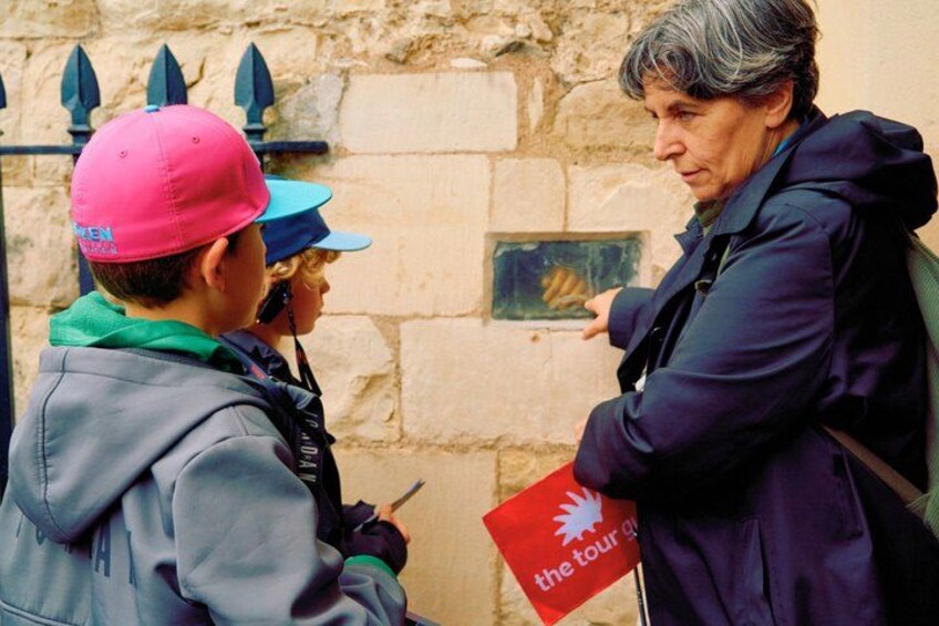 Una persona con una chaqueta oscura sostiene un letrero rojo con un logotipo, de pie frente a un muro de piedra con una reja de metal. 3