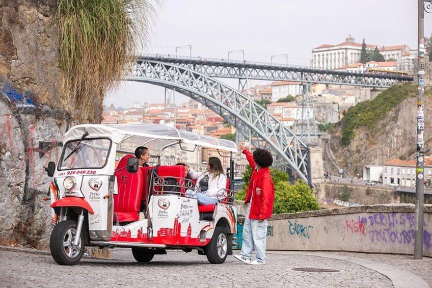 A white tuk-tuk with red accents parked near a bridge, a person in red standing nearby, and a cityscape in the background. 2