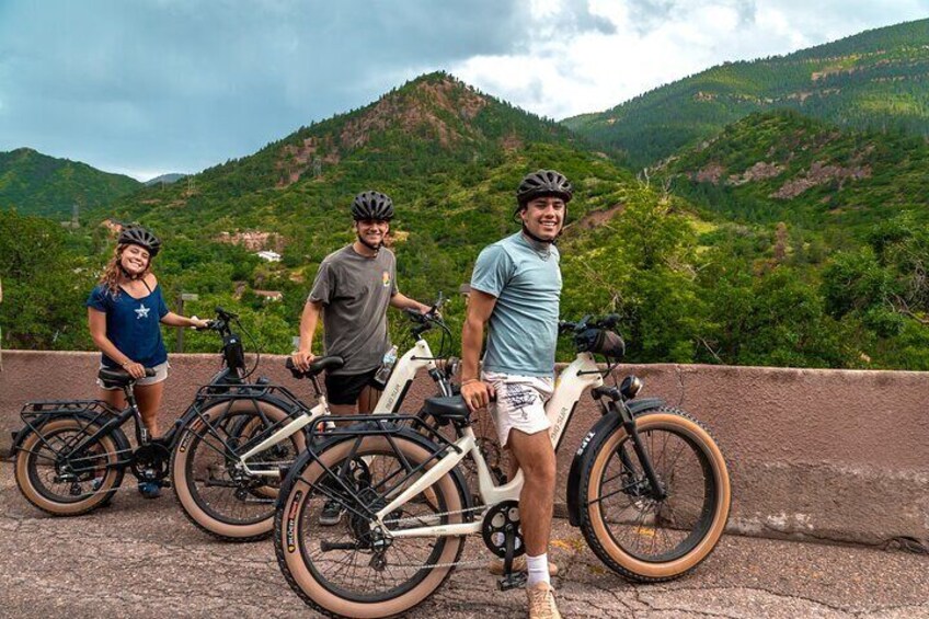 Three people standing with bicycles on a bridge with a mountainous landscape in the background. 3