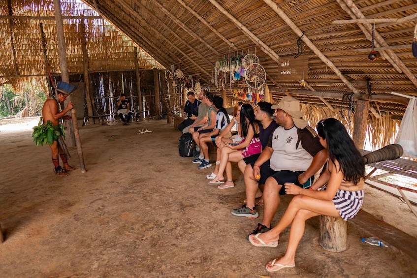 A group of people sitting on benches in a thatched-roof hut with hanging decorations. 2
