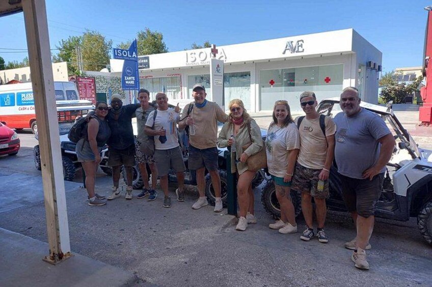 A group of people posing in front of a building with 'ISOLA' signage, a white vehicle, and a red car in the background. 3