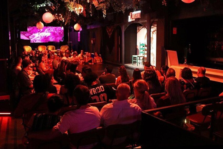 An indoor event with an audience seated in rows, a stage with a performer, and a large screen displaying visuals. 2
