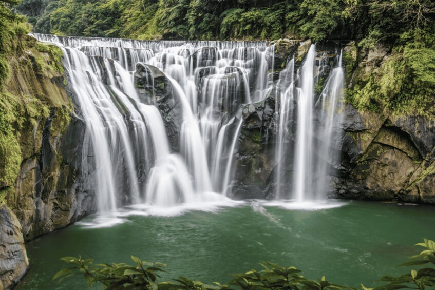 A multi-tiered waterfall cascading into a green pool surrounded by rocky cliffs and lush greenery. 2