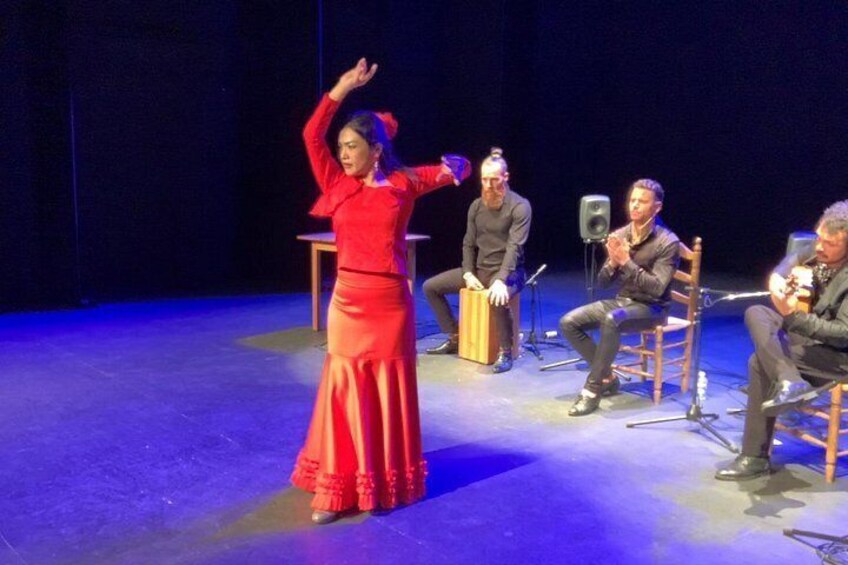 A person in a red dress performing on stage with a microphone stand and audience seated on chairs. 4