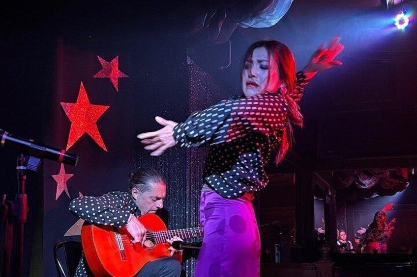 A person playing a red guitar on stage with a microphone stand, a star decoration, and another person in the background. 5