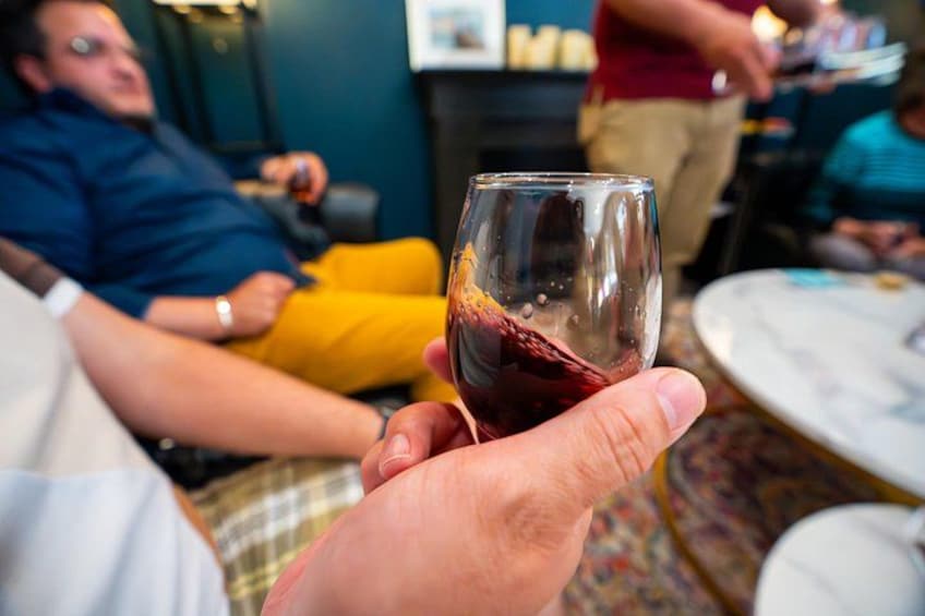 Una persona sosteniendo una copa de vino con un líquido oscuro, posiblemente vino tinto, en un ambiente social con otras personas al fondo. 4