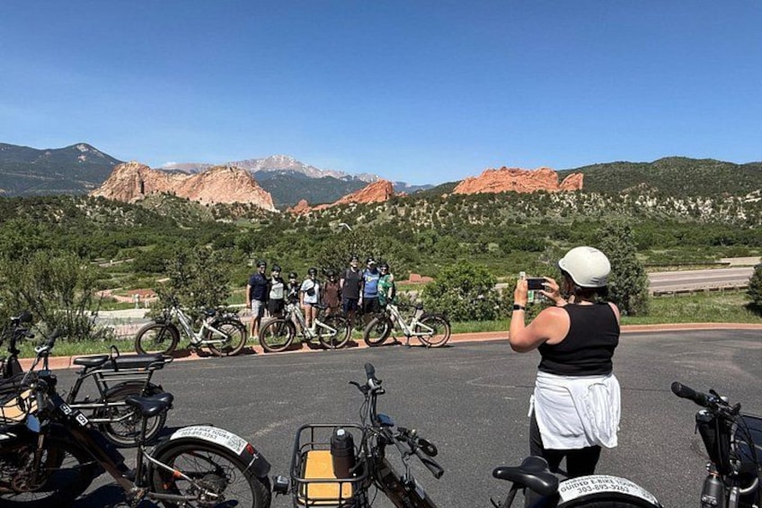 A group of cyclists and a person taking a photo in a parking lot with bicycles, mountains, and red rock formations in the background. 4