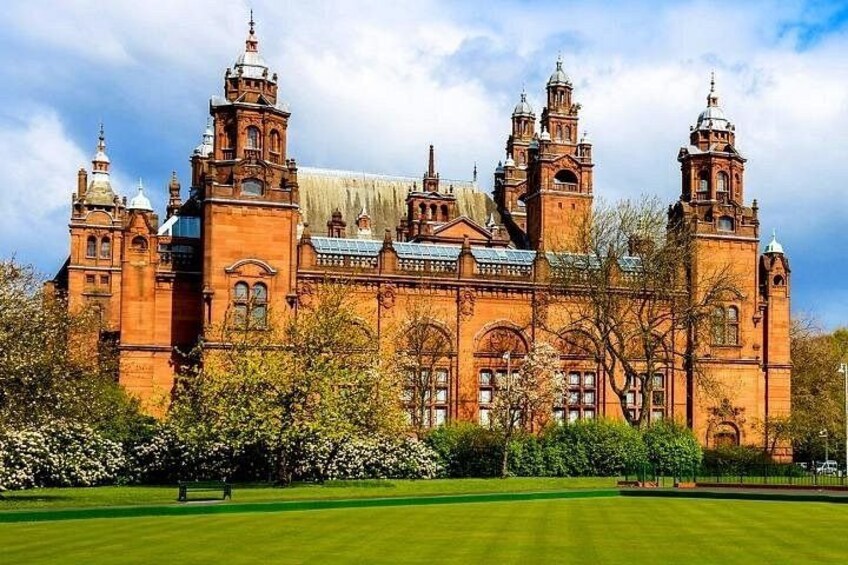 A large, historic building with multiple towers and arched windows, surrounded by a well-maintained lawn and flowering trees. 1