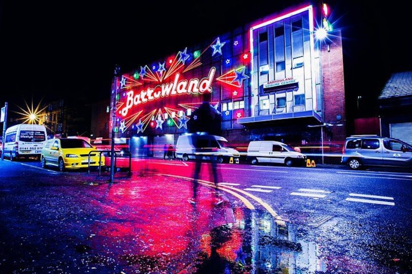 A brightly lit building with a neon sign reading 'Battleground' and a reflection on wet pavement. 5