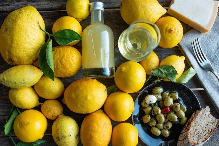 Limoni, una bottiglia di olio d'oliva, un bicchiere di vino bianco, un piatto di olive e una fetta di pane su un piano di legno. 2