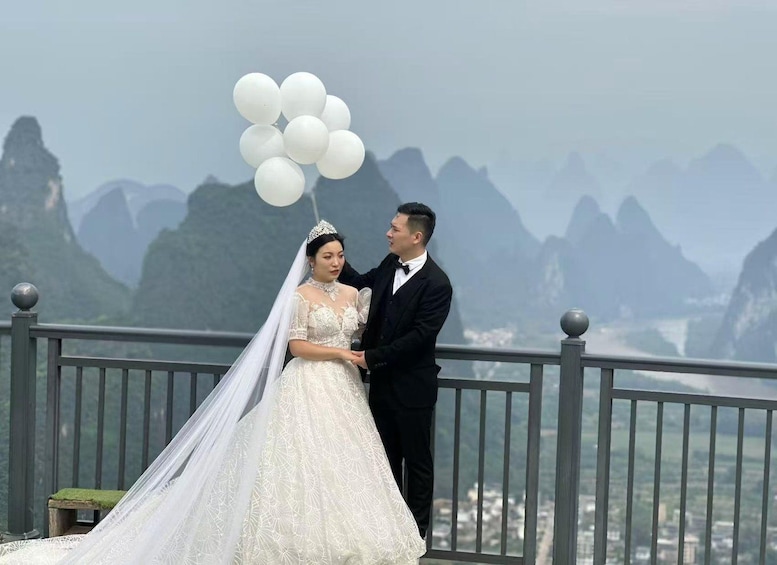 A bride and groom holding white balloons, standing on a balcony with a mountainous landscape in the background. 4