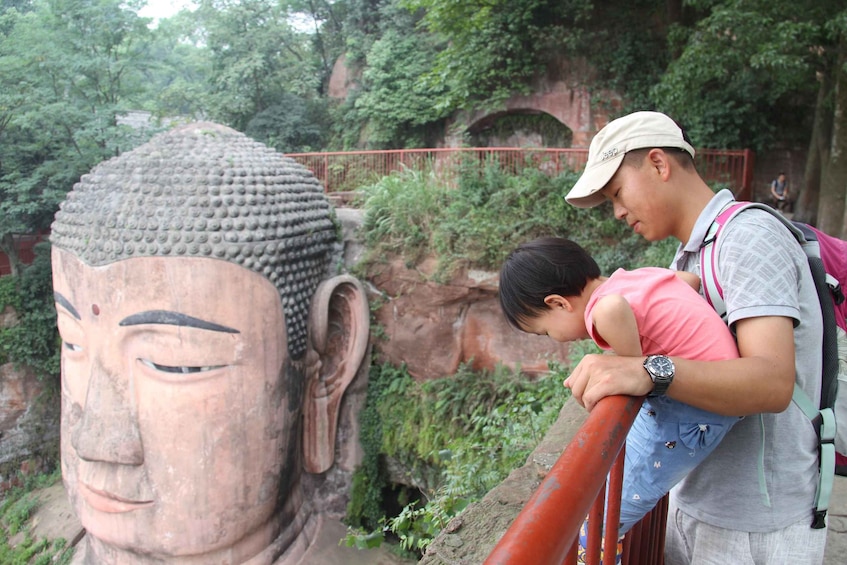 A large stone Buddha statue, a person leaning on a railing, and a red fence in a natural setting. 3