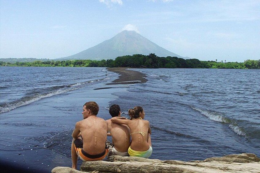 Three people sitting on a rocky shore, facing a large volcano in the distance. 1
