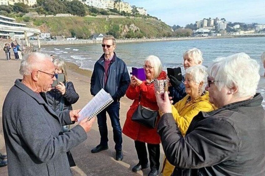Eine Gruppe von Menschen steht an einer Promenade am Meer, im Hintergrund sind Gebäude zu sehen. 3