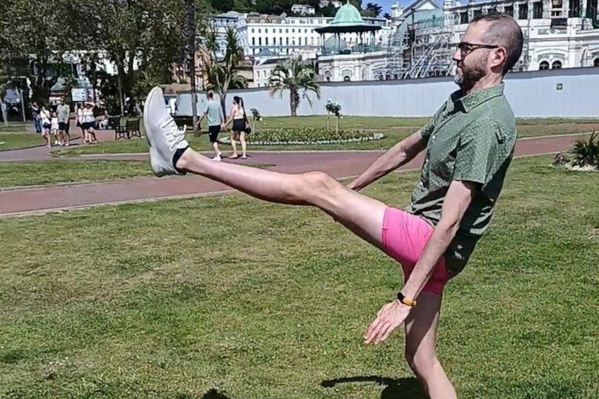 A person performing a leg stretch outdoors with buildings and a park in the background. 3