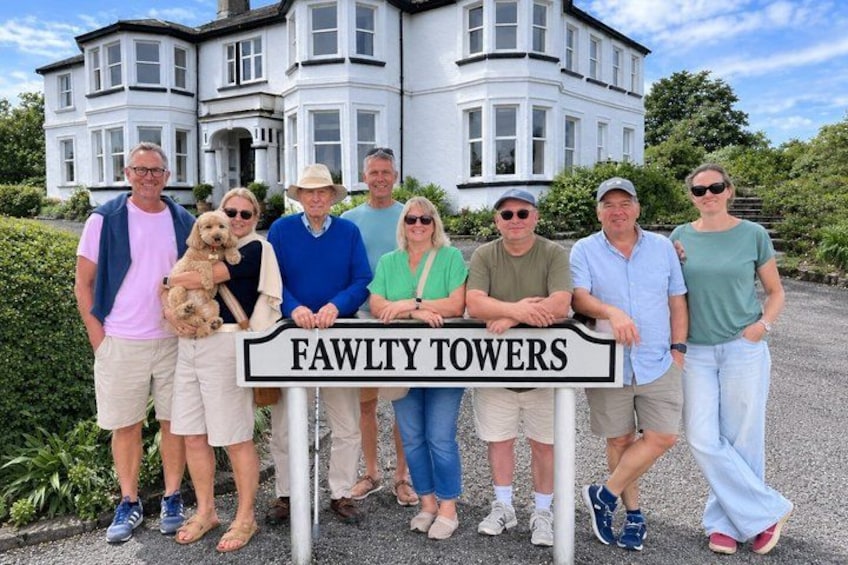 A group of people standing in front of a sign that reads 'FAWLTY TOWERS' with a large house in the background. 2