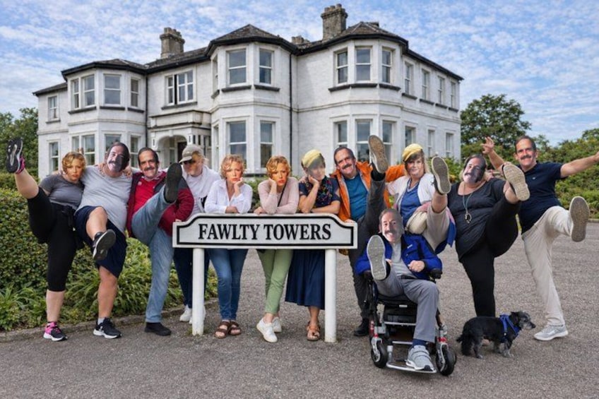A group of people posing in front of a sign that reads 'FAWLTY TOWERS' with a large white building in the background. 1